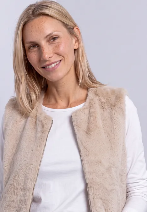 Lächelnde Frau mit blonden Haaren trägt weißes Langarmshirt und beigen Pelzimitat-Weste vor grauem Hintergrund