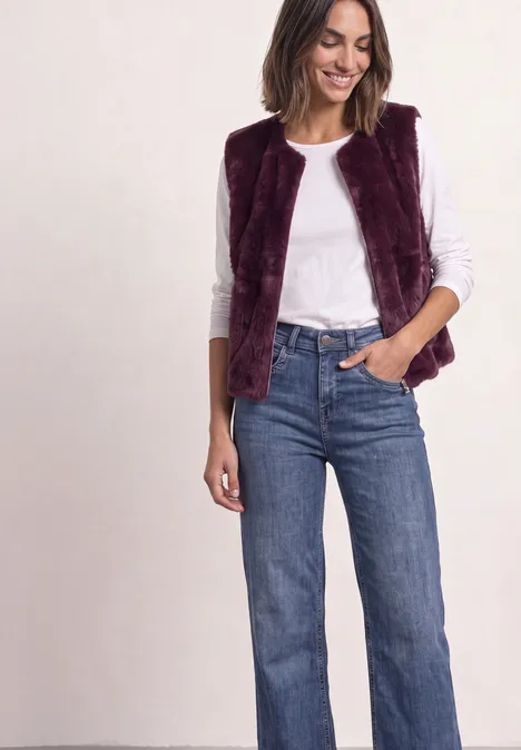 Frau in weißem Langarmshirt, burgunderroter, pelziger Weste und weiten blauen Jeans.