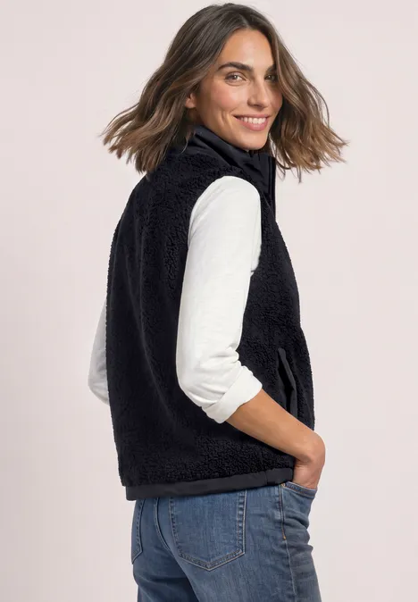 Eine Frau trägt eine schwarze Fleeceweste über weißem Shirt und Jeans, schaut seitlich zur Kamera.