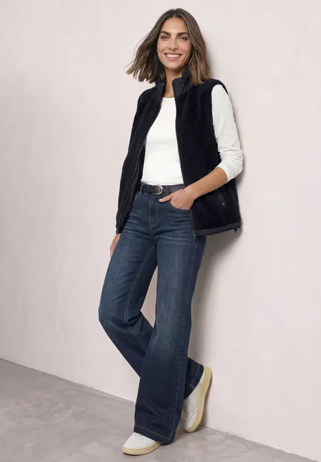 Eine Frau in einer schwarzen ärmellosen Weste, weißem Shirt und weiten Jeans vor hellem Hintergrund.