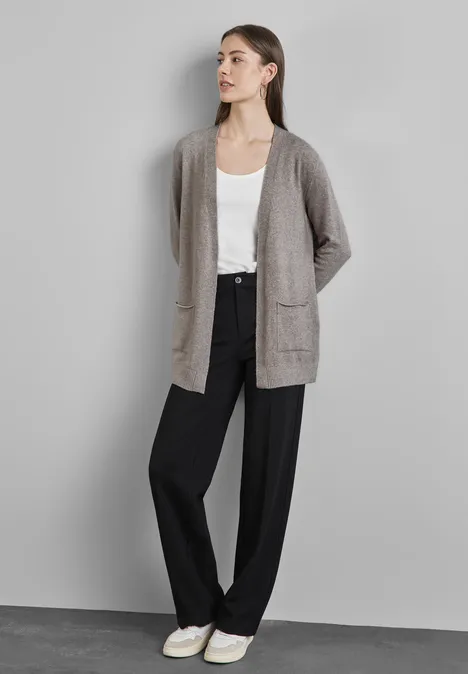 Frau mit grauer Strickjacke, weißem Top und schwarzer Hose steht vor einer grauen Wand.