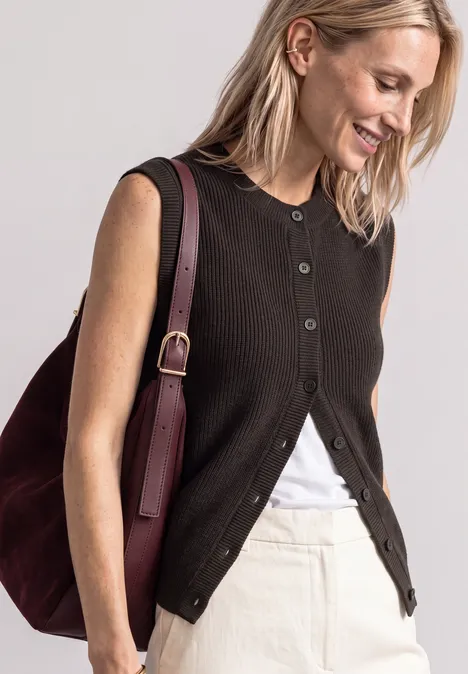 Een glimlachende vrouw draagt een zwart mouwloos vest, een wit topje, een lichte rok en een bordeaux tas.