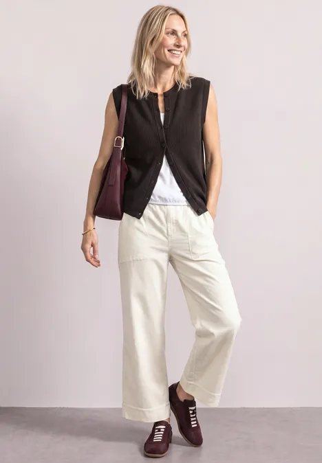 Blonde vrouw met zwart gilet, wijde witte broek, bordeaux schoenen en tas, tegen een lichte achtergrond.