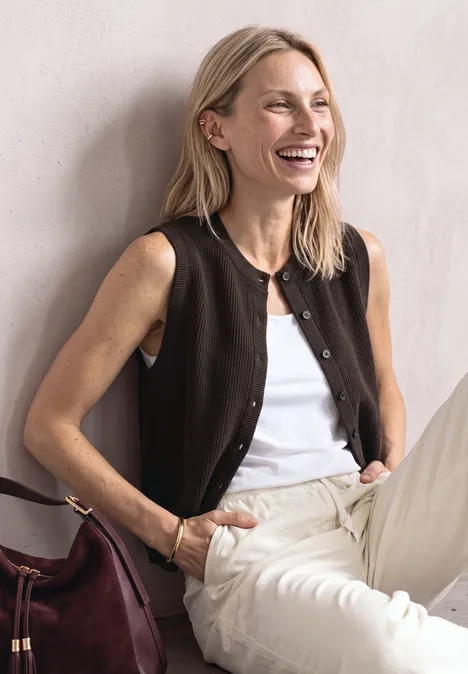 Een glimlachende blonde vrouw zit voor een lichte muur, draagt een wit topje, bruin vest, lichte broek en een bordeaux tas.