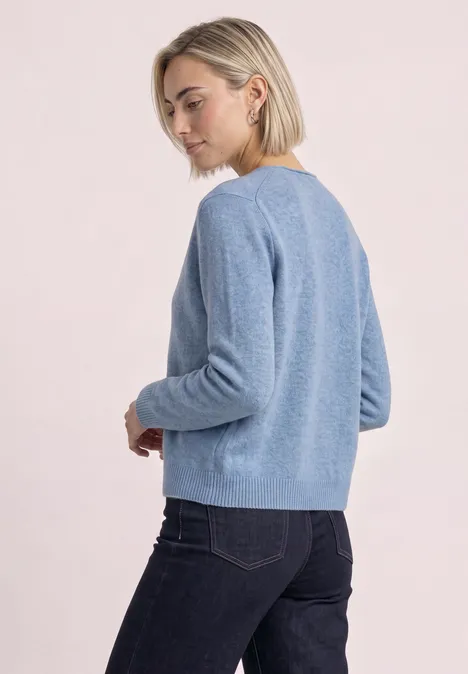 Cardigan à col en V et patte de boutonnage northern blue mel. Une femme porte un pull bleu clair et un jean foncé, posant de côté et regardant par-dessus son épaule.