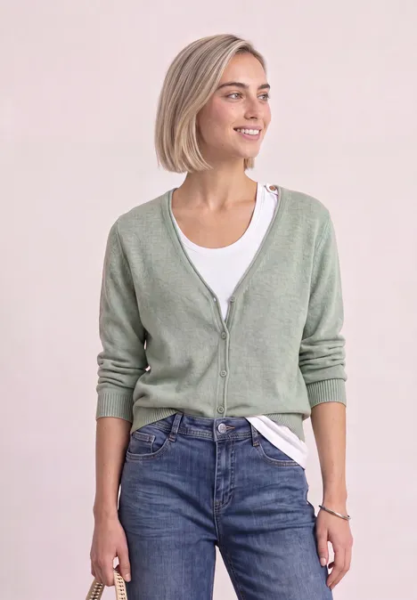 Cardigan à col en V et patte de boutonnage eucalyptus green mel. Jeune femme aux cheveux blonds courts porte un gilet vert pâle, tee-shirt blanc et jean, sourit.