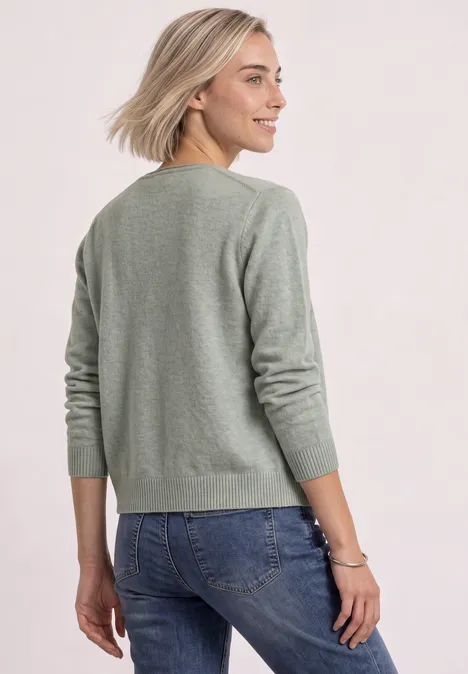 Cardigan à col en V et patte de boutonnage eucalyptus green mel. Femme de dos portant un pull vert et un jean bleu, cheveux blonds, sur fond clair.