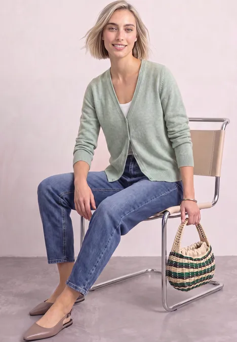 Cardigan à col en V et patte de boutonnage eucalyptus green mel. Femme en cardigan vert, jean et ballerines beiges, assise sur une chaise, tenant un sac tressé.
