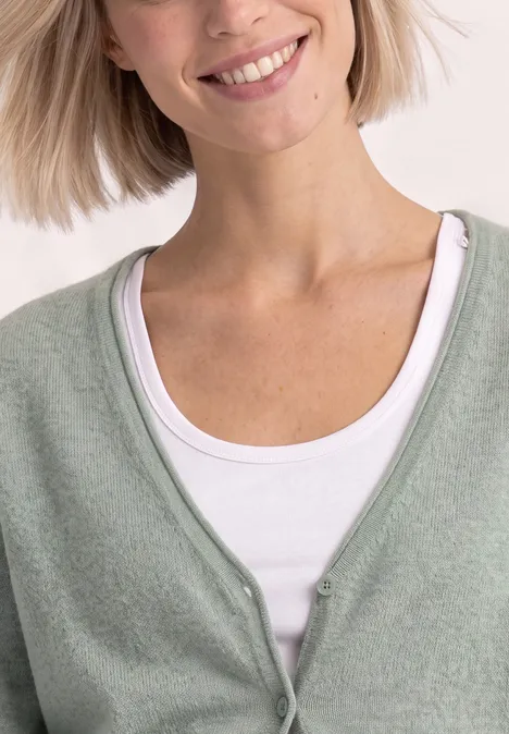 Cardigan à col en V et patte de boutonnage eucalyptus green mel. Femme souriante portant un gilet vert clair sur un top blanc.