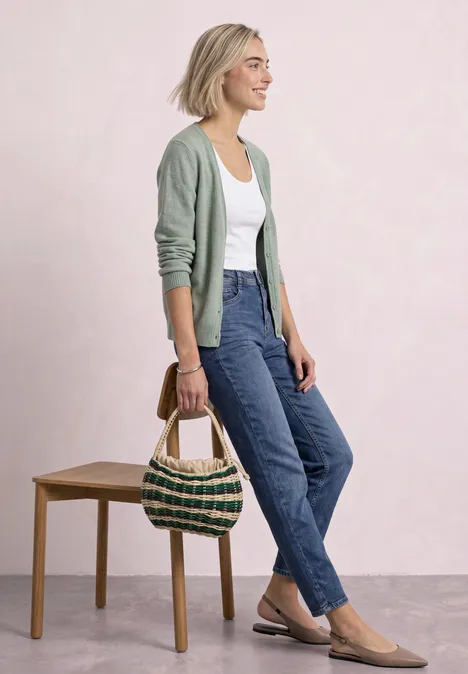 Cardigan à col en V et patte de boutonnage eucalyptus green mel. Femme en gilet vert, débardeur blanc, jean bleu et sac en paille sur fond clair.
