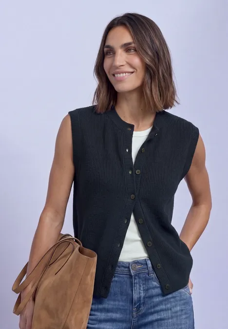 Vrouw in zwarte mouwloze gilet over een wit shirt, glimlachend met een bruine tas in de hand.