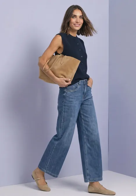 Vrouw in zwart mouwloos topje, wijd blauwe jeans, beige loafers en grote bruine tas voor een lichte achtergrond.