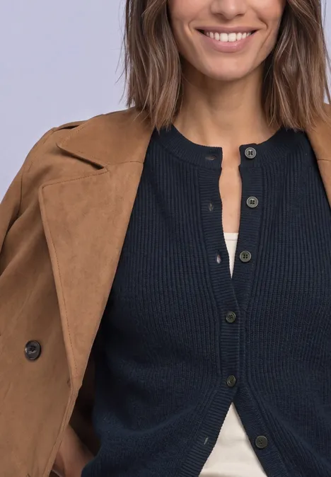 Een glimlachende vrouw met een bruine jas over haar schouders en een zwarte gebreide cardigan.