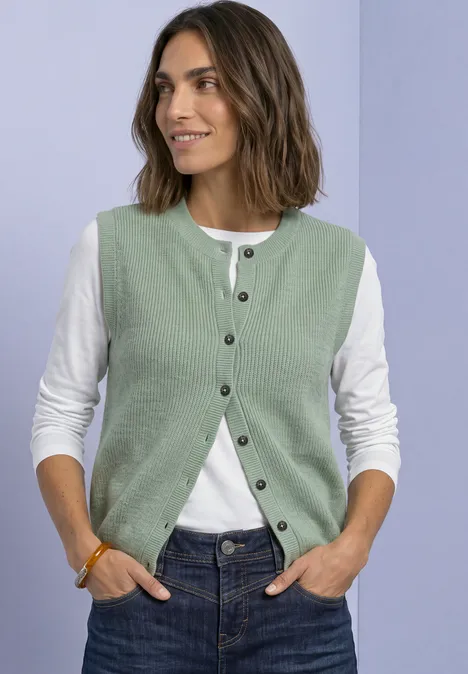 Strickweste mit Rundhals und Knopfleiste eucalyptus green mel. Frau in grüner Strickweste über weißem Langarmshirt und Jeans, lächelnd, Hände in den Taschen.