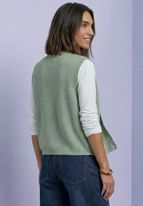 Strickweste mit Rundhals und Knopfleiste eucalyptus green mel. Eine Frau trägt eine grüne Strickweste über weißem Langarmshirt und Jeans vor lila Hintergrund.