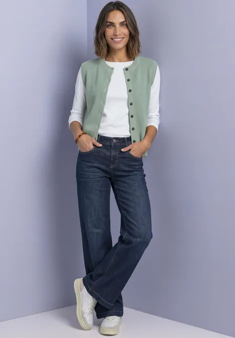 Strickweste mit Rundhals und Knopfleiste eucalyptus green mel. Frau mit grün-weißer Strickjacke, weißem Shirt, blauen Jeans und weißen Sneakern vor fliederfarbenem Hintergrund.