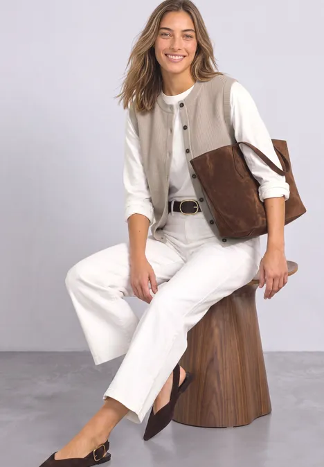 Een lachende vrouw zit op een houten kruk, draagt witte kleding en een bruine leren tas.