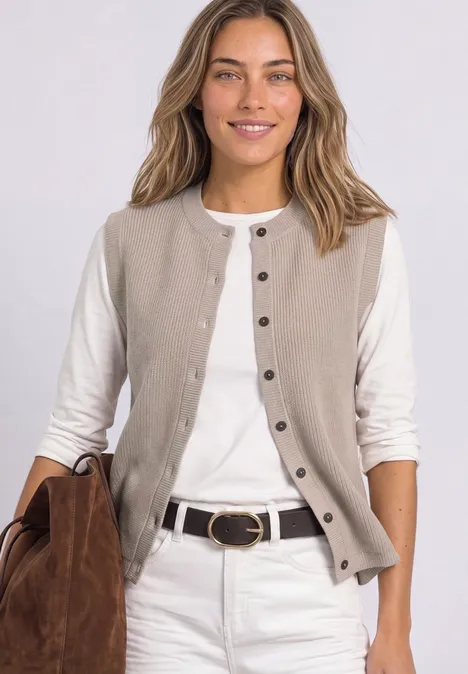 Vrouw lacht, draagt beige mouwloos vest, wit shirt, witte jeans en een bruine tas.