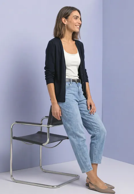 Cardigan à col en V et patte de boutonnage darkest blue Femme en jean, débardeur blanc et cardigan, appuyée sur une chaise moderne devant un fond pastel.