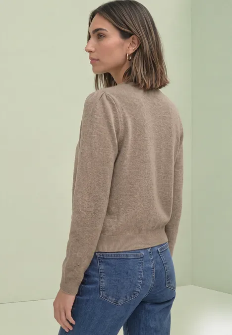 Frau mit kurzem Haar in beigem Pullover und blauen Jeans steht seitlich vor hellem Hintergrund.