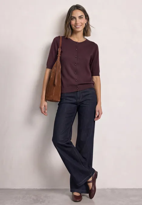 Kurzarm Cardigan mit Rundhals und Knöpfen rustic rouge Frau in lila Oberteil und ausgestellten Jeans, trägt Tasche und locker lächelnd.