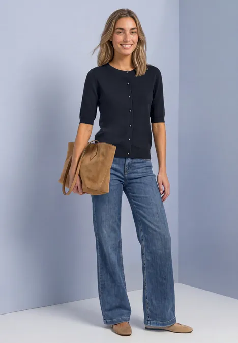 Vrouw lacht, draagt een donkerblauw vest, wijdvallende jeans, comfortabele schoenen en een bruine tas over de schouder.