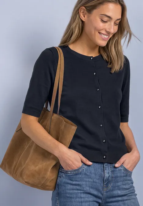 Een glimlachende vrouw draagt een zwarte cardigan, spijkerbroek en een bruine suède shopper.