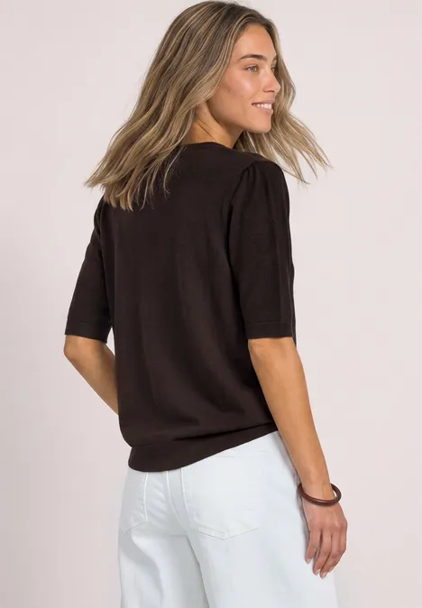 Jonge vrouw in zwarte korte mouw trui en witte broek, zijaanzicht van achteren, glimlachend voor lichte achtergrond.
