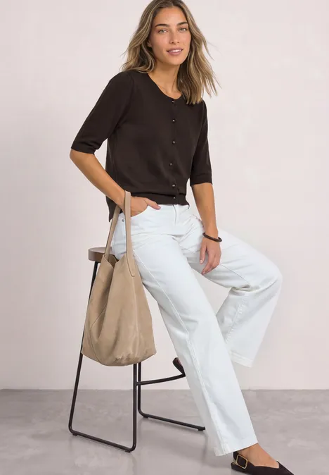 Vrouw in donker shirt en witte jeans met canvas tas, zittend op een kruk voor een lichte achtergrond.