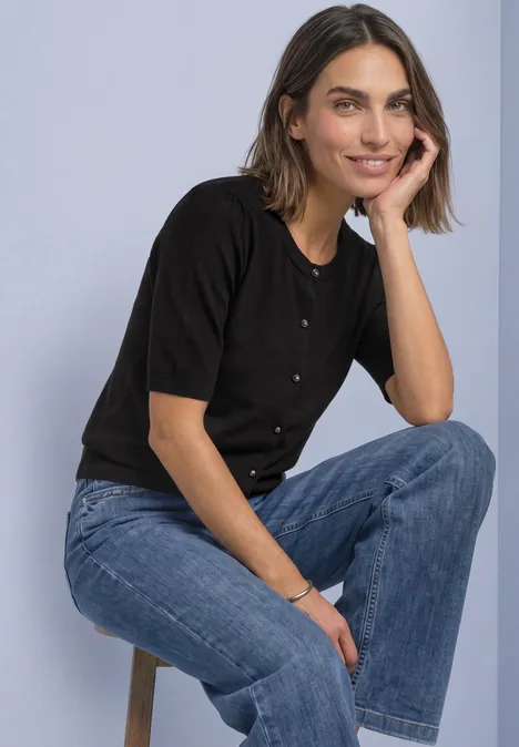 Een glimlachende vrouw in een zwart shirt en blauwe jeans zit ontspannen op een krukje.