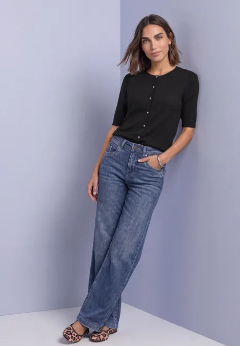 Vrouw leunt nonchalant tegen wand, draagt zwart vest, wijde jeans en luipaardprint sandalen voor lichte achtergrond.