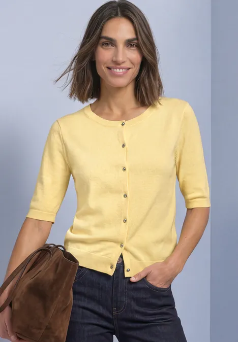 Vrouw met gele korte mouw cardigan met knopen, donkere jeans en bruine schoudertas.