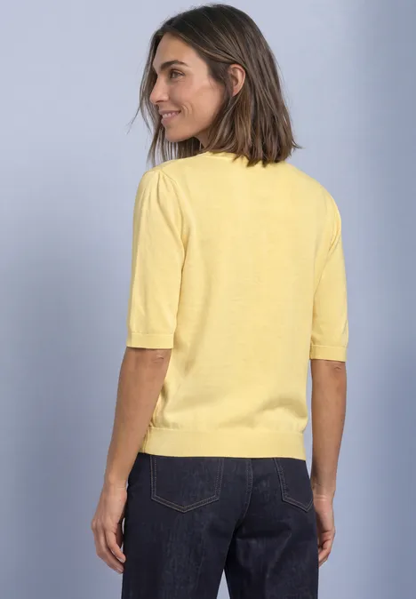 Vrouw van achteren in een gele top met korte mouwen en een donkere spijkerbroek tegen neutrale achtergrond.