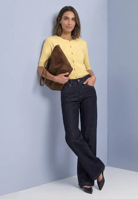 Vrouw in geel shirt en donkerblauwe jeans, met een bruine tas onder de arm, leunend tegen een muur.
