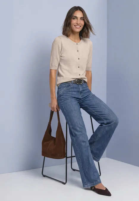 Vrouw leunt op stoel, glimlacht, draagt licht truitje, wijde jeans, bruine tas en puntige schoenen.