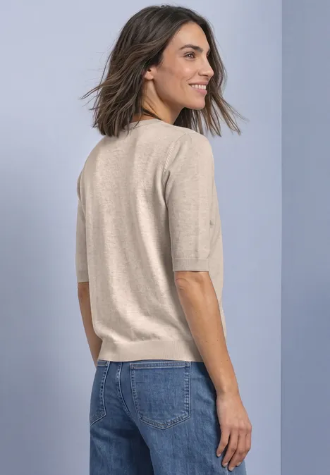 Vrouw van achter, halflang bruin haar, draagt een beige trui met korte mouwen en een blauwe jeans, kijkt over haar schouder.