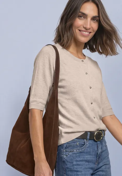 Glimlachende vrouw in spijkerbroek, beige kort vest en grote bruine schoudertas voor een lichte achtergrond.