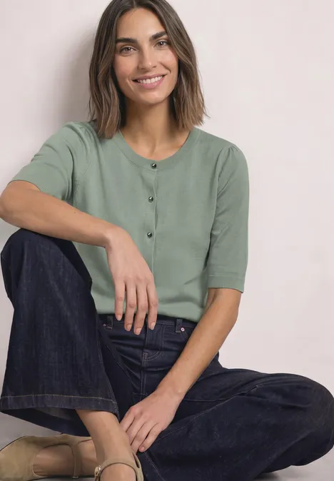 Vrouw zit glimlachend, draagt een groen shirt en een donkere spijkerbroek.