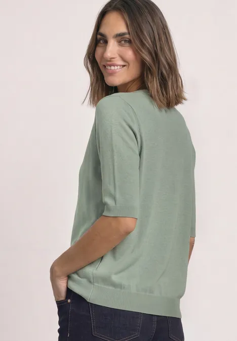 Vrouw in een groene trui met korte mouwen en donkere jeans kijkt lachend over haar schouder.