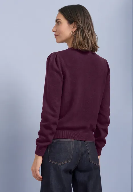 Kardigan z długimi rękawami, okrągłym dekoltem i guzikami berry maroon mel. Kobieta w profilu ma na sobie bordowy sweter i ciemne jeansy na neutralnym tle.