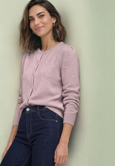 Eine lächelnde Frau in rosa Strickjacke und dunkler Jeans lehnt leicht an einer Wand.