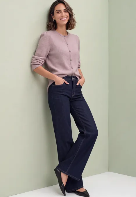 Eine lächelnde Frau lehnt entspannt an einer grünen Wand, trägt rosa Cardigan und dunkelblaue Jeans.