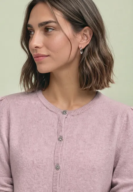Junge Frau mit kurzen braunen Haaren und rosa Strickjacke blickt seitlich, dezentes Make-up und zierliche Ohrringe.