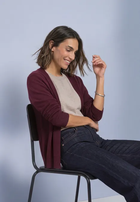 Langarm Cardigan im offenen Schnitt rustic rouge Eine lächelnde Frau sitzt auf einem Stuhl, trägt einen weinroten Cardigan, beige Top und blaue Jeans.