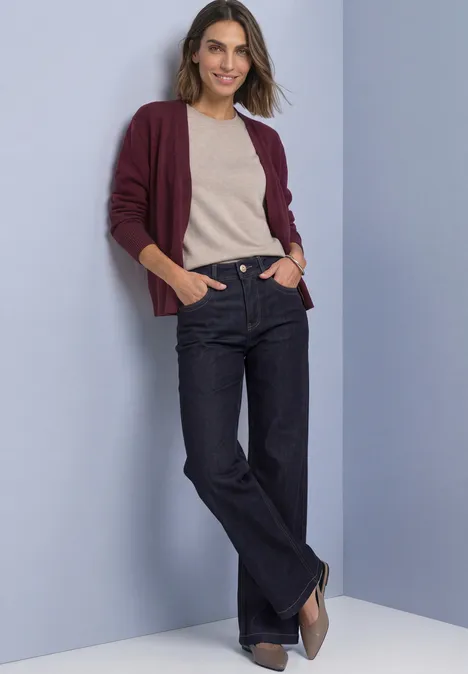Langarm Cardigan im offenen Schnitt rustic rouge Frau lehnt lässig vor pastellfarbenem Hintergrund, trägt weinrote Strickjacke, beiges Top, dunkle Jeans und Pumps.