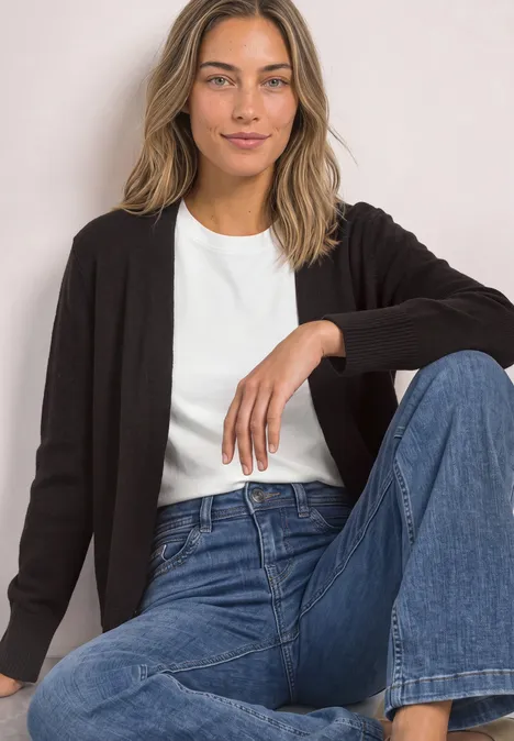 Langarm Cardigan im offenen Schnitt black coffee Frau sitzt, trägt weiße T-Shirt, schwarze Strickjacke und blaue Jeans, posiert vor hellem Hintergrund.