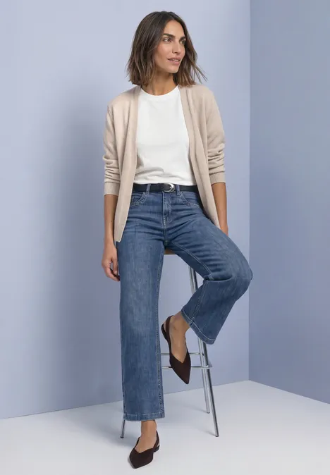Femme portant un t-shirt blanc, un cardigan beige, un jean droit bleu et des ballerines brunes devant un mur clair.