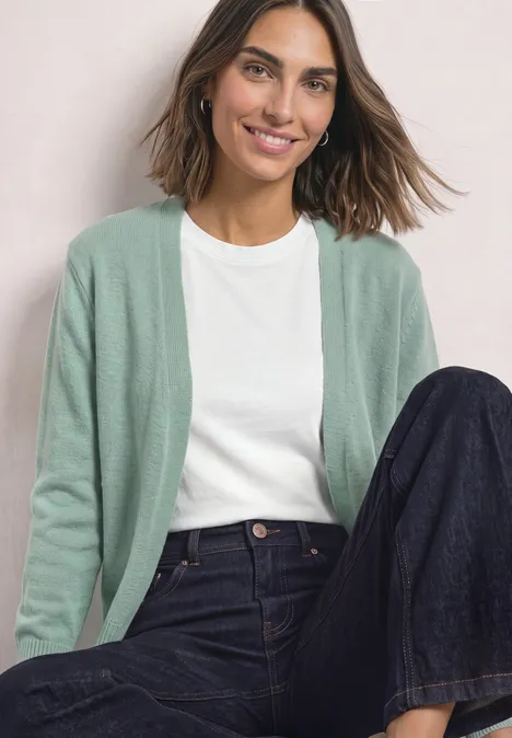 Femme assise souriante, portant un gilet vert, un t-shirt blanc et un jean ample foncé.
