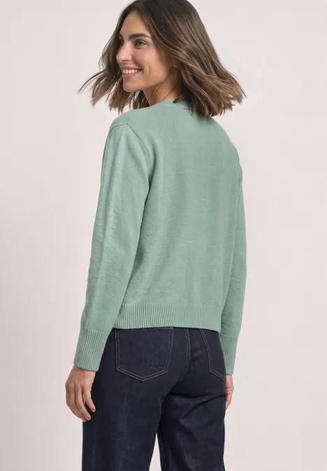 Une femme porte un pull vert clair et un jean foncé, souriante, regard par-dessus son épaule.