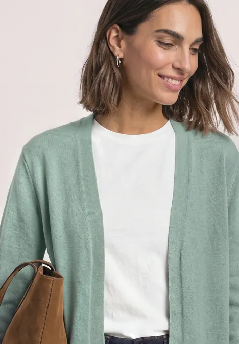 Femme souriante en t-shirt blanc et cardigan vert menthe, avec un sac marron à l'épaule.